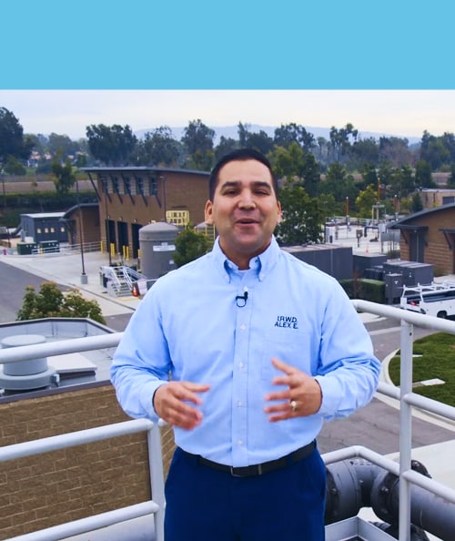 Man in a light blue button-down shirt with a microphone on a rooftop overlooking campus buildings.