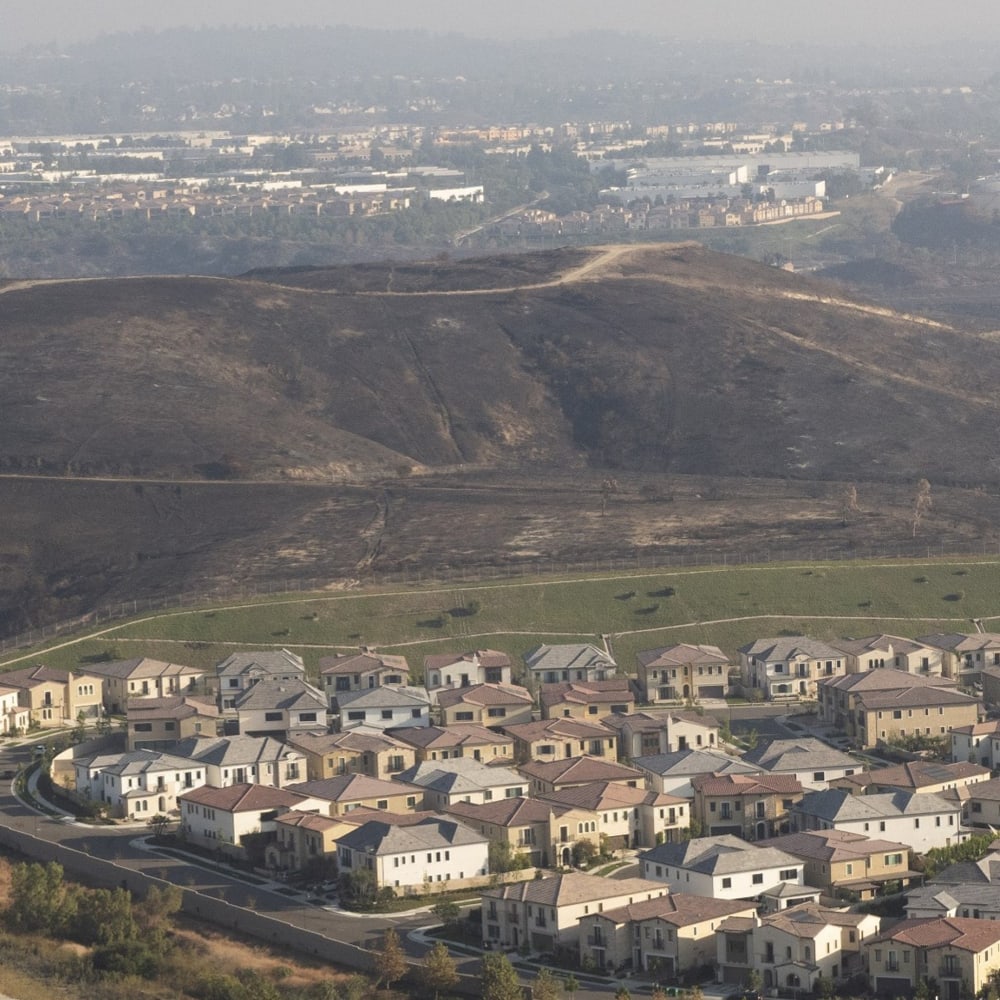 Suburban neighborhood at the base of a dry, rolling hill with distant industrial buildings on the horizon