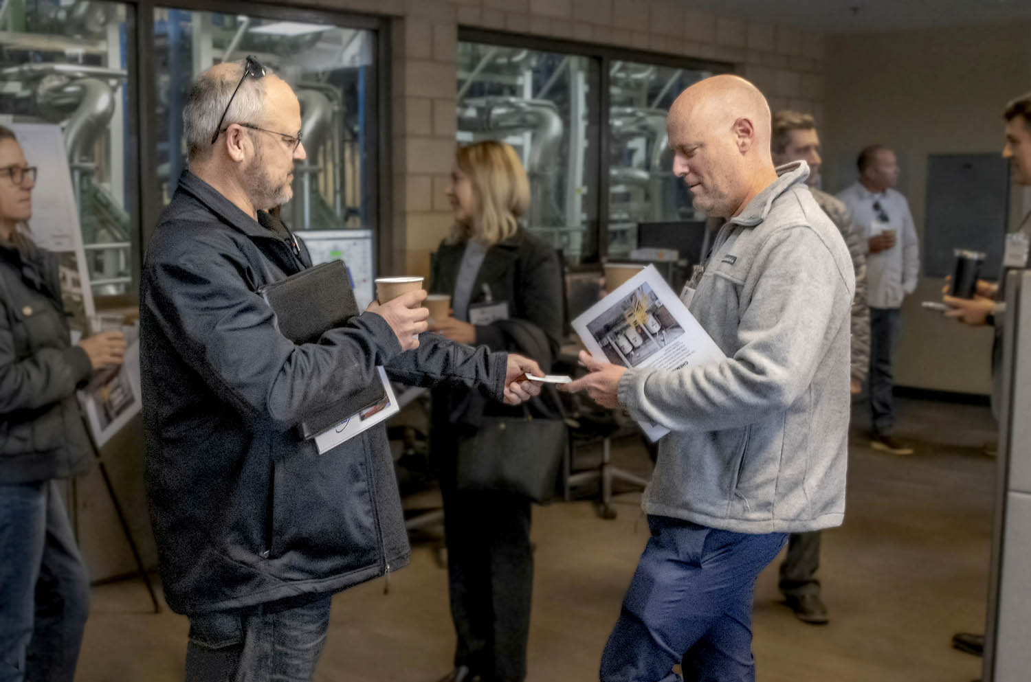 Two men exchange business cards in a facility lobby while coworkers chat in the background.