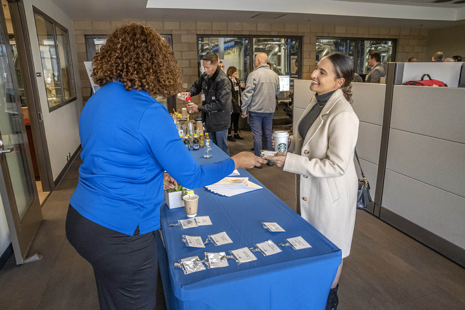 Woman in a beige coat hands ID badge to attendee at a blue-table check-in station during an office event.