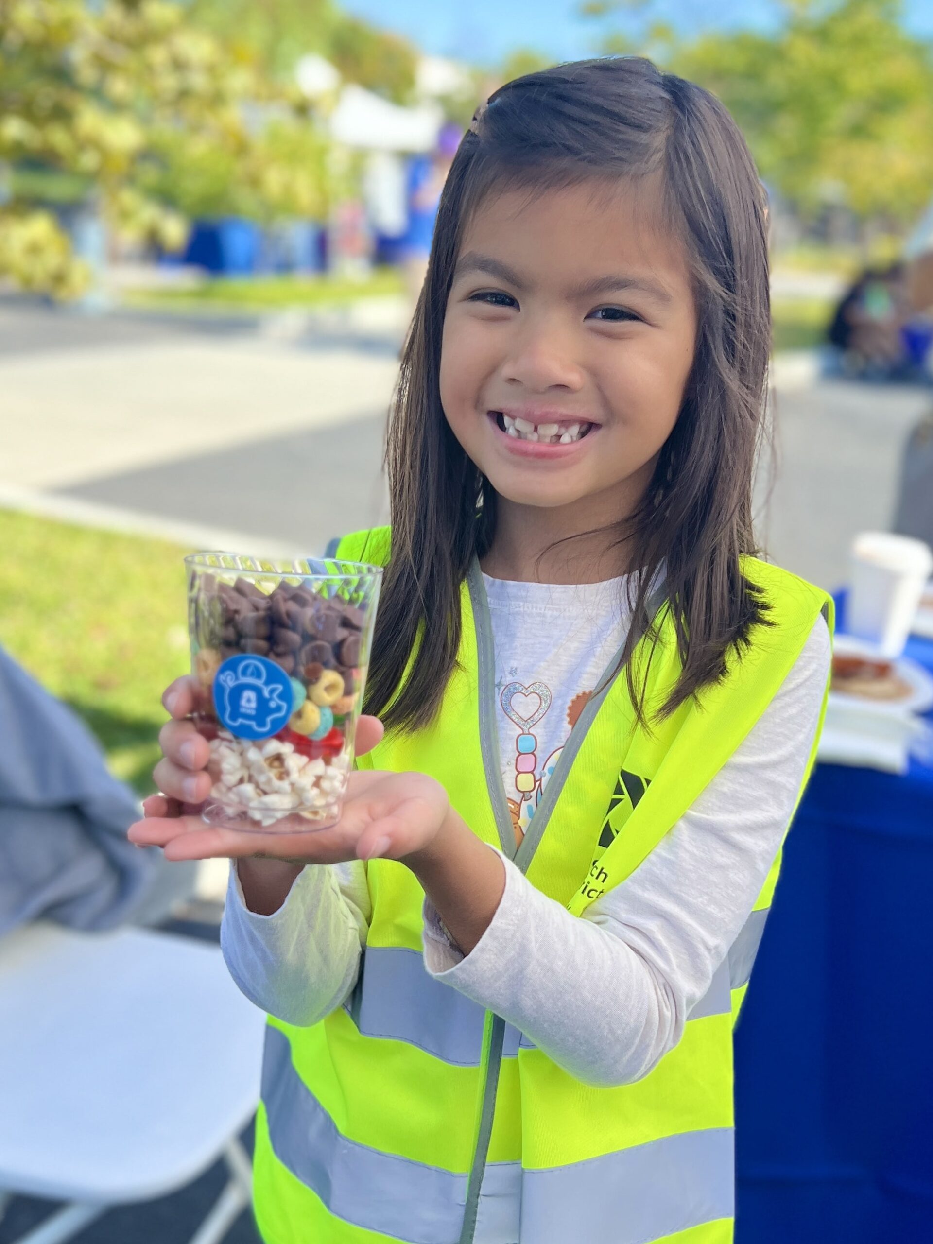 Smiling girl in a neon safety vest holds a clear cup of mixed snacks at an outdoor event.