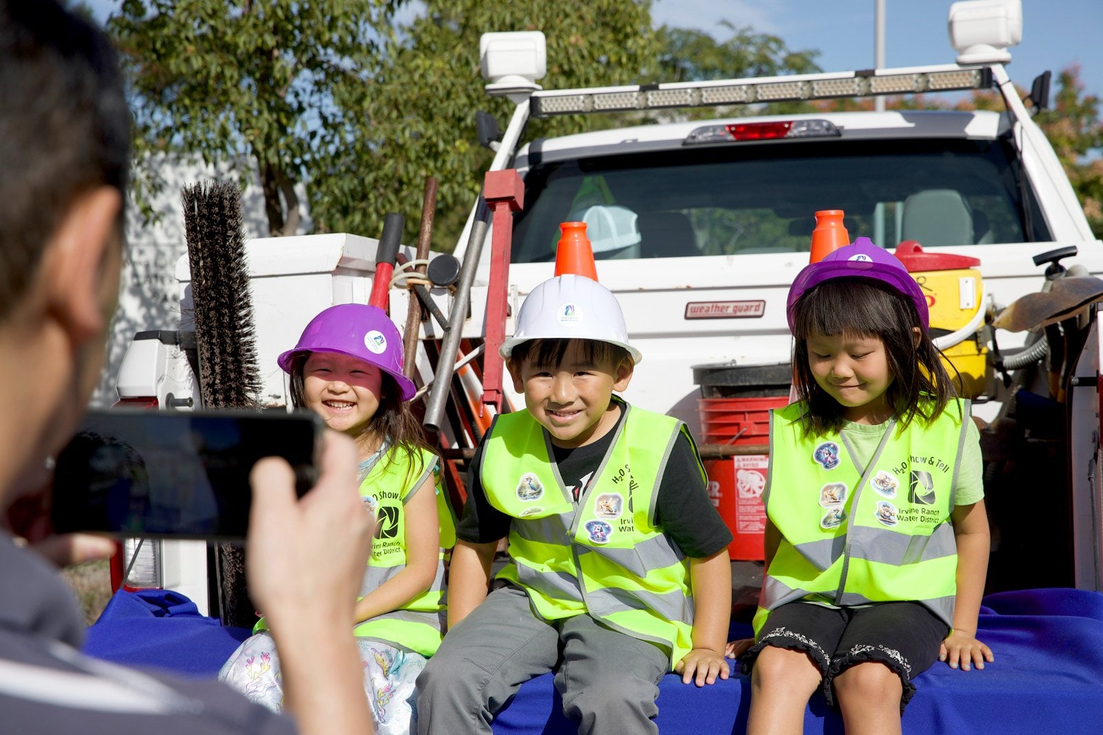 Three children wearing neon safety vests and hard hats sit in front of a white utility truck during a safety event.