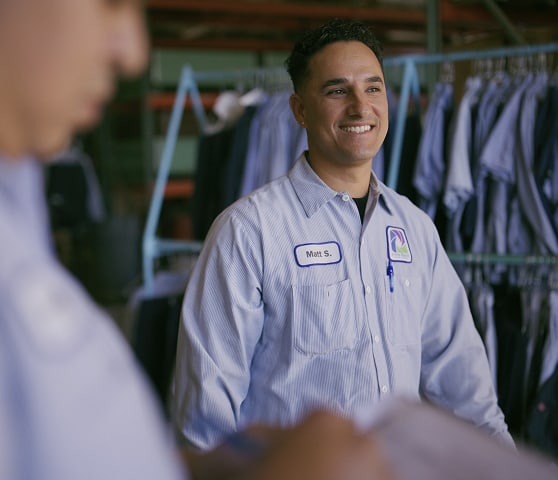 Smiling male staff member in a light-blue striped uniform with a visible badge, standing near clothing racks.