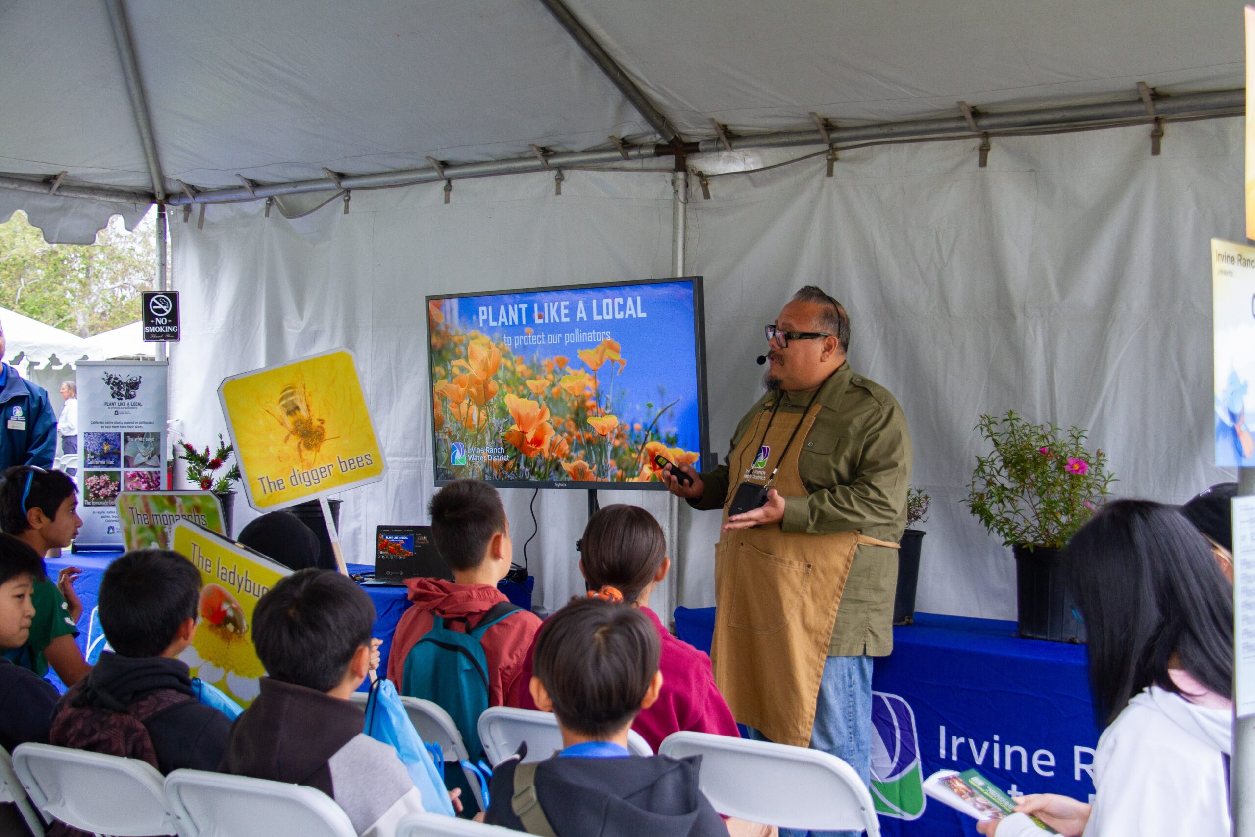 Speaker in a green shirt and apron talks to kids in a tented booth, presenting a 'Plant Like a Local' display on a large screen.