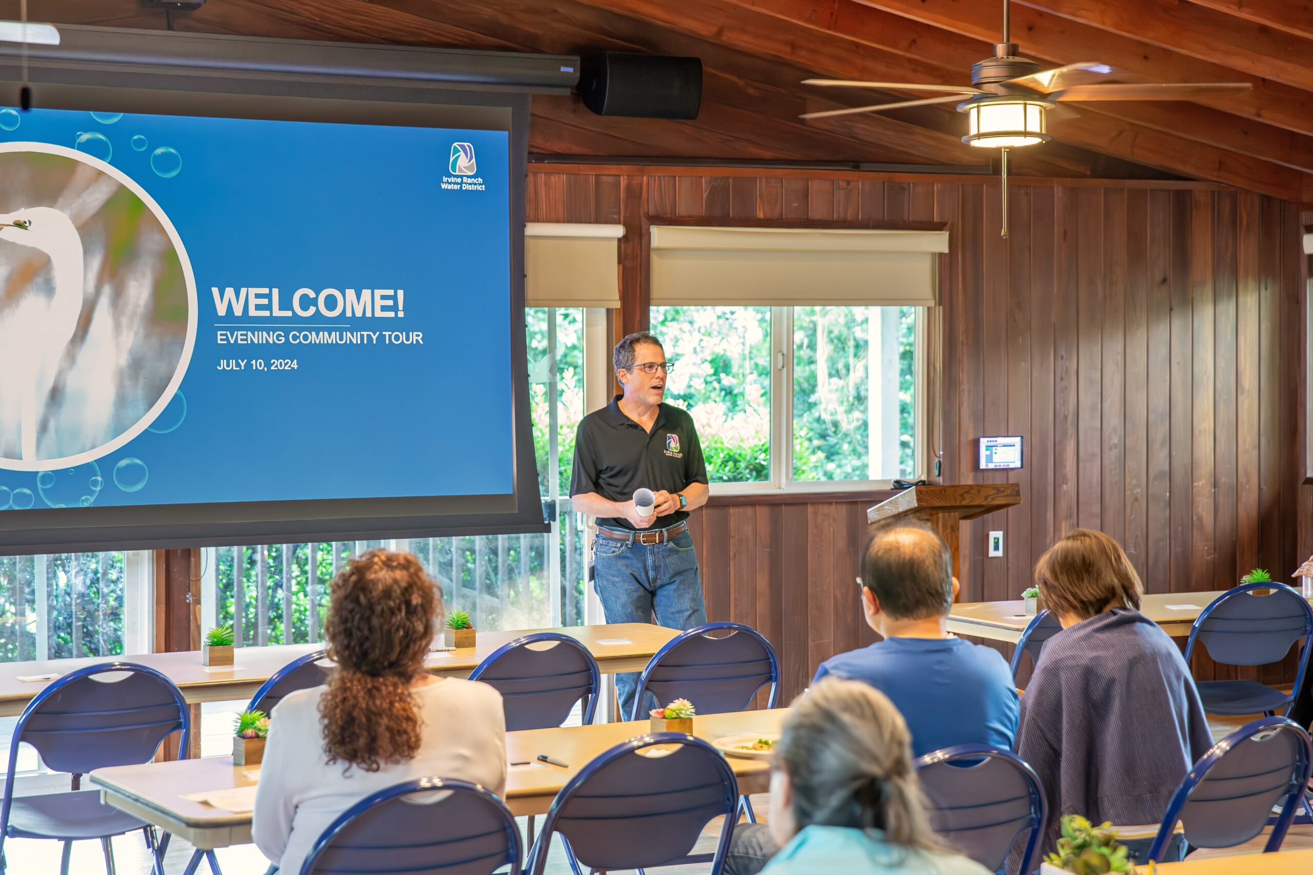 Speaker in a black polo at a podium presenting to an audience in a wood-paneled room beside a blue Welcome slide.