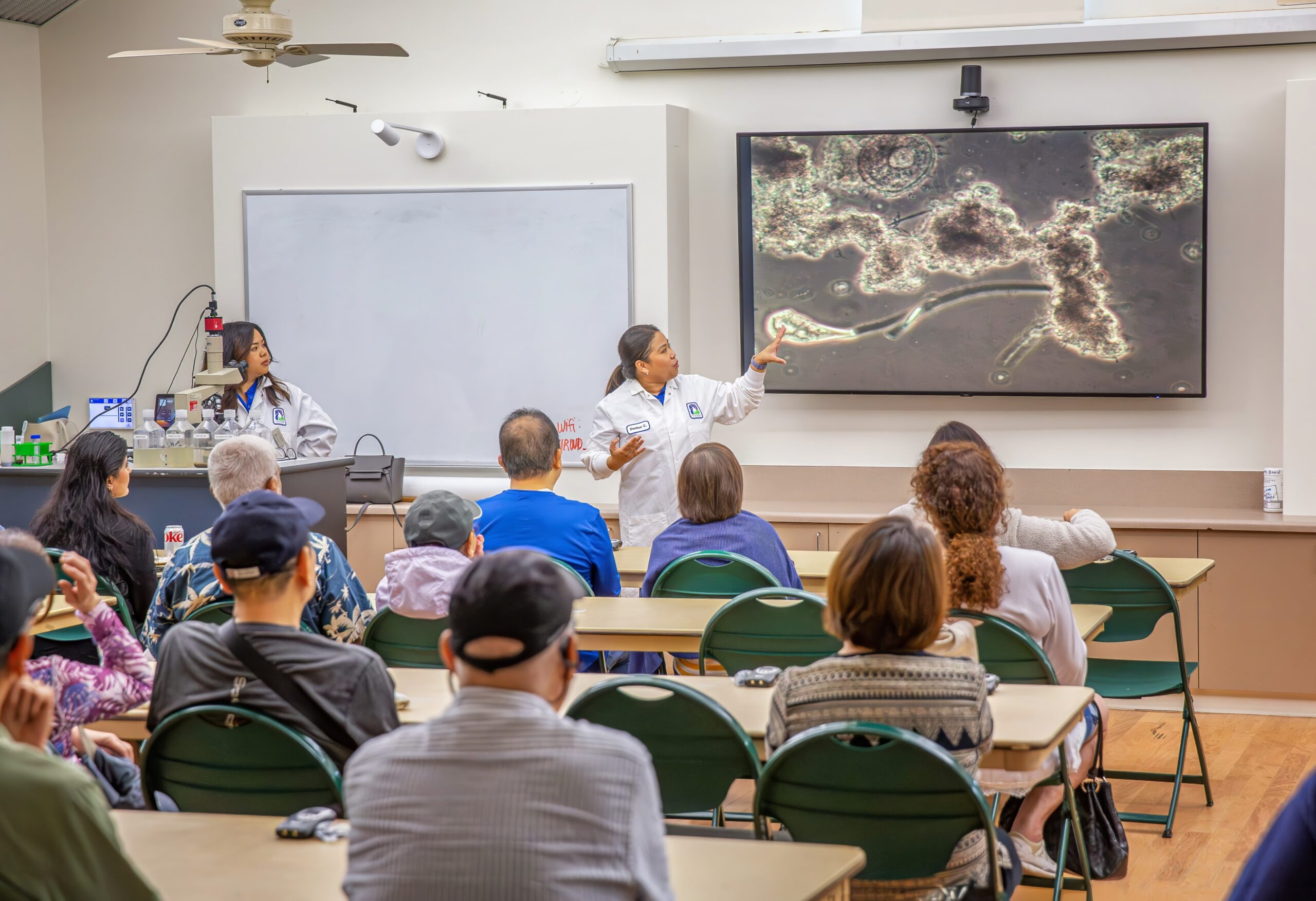 Instructor in a white lab coat points to a screen showing magnified cellular structures as students watch in a classroom lab.