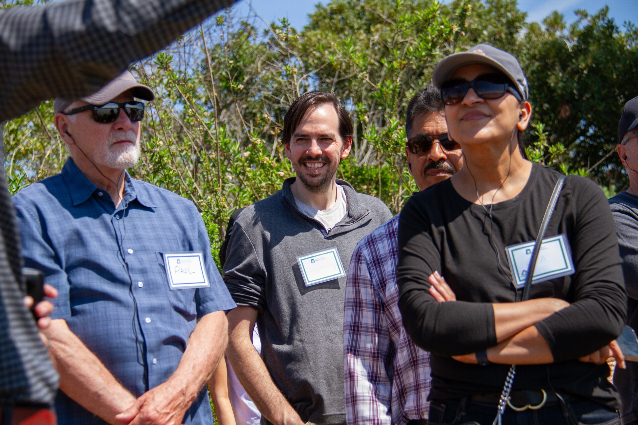 Group of adults wearing name badges at an outdoor daytime gathering, with trees in the background.
