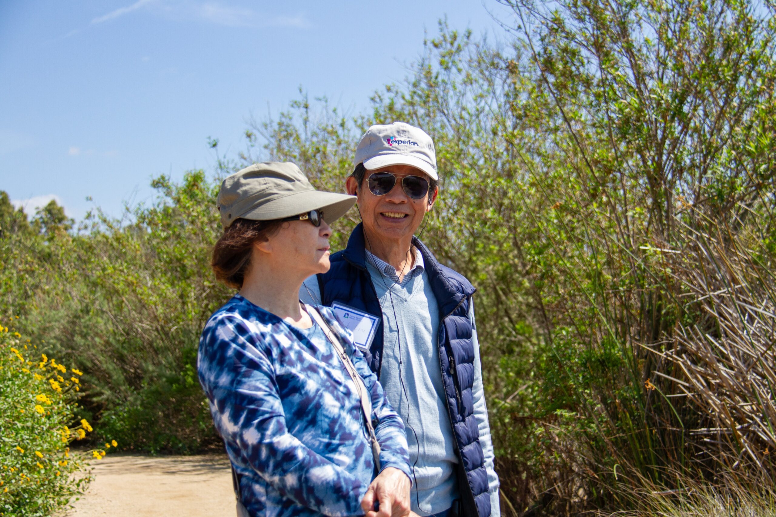 Two hikers wearing hats and sunglasses walk along a sunny nature trail surrounded by shrubs.