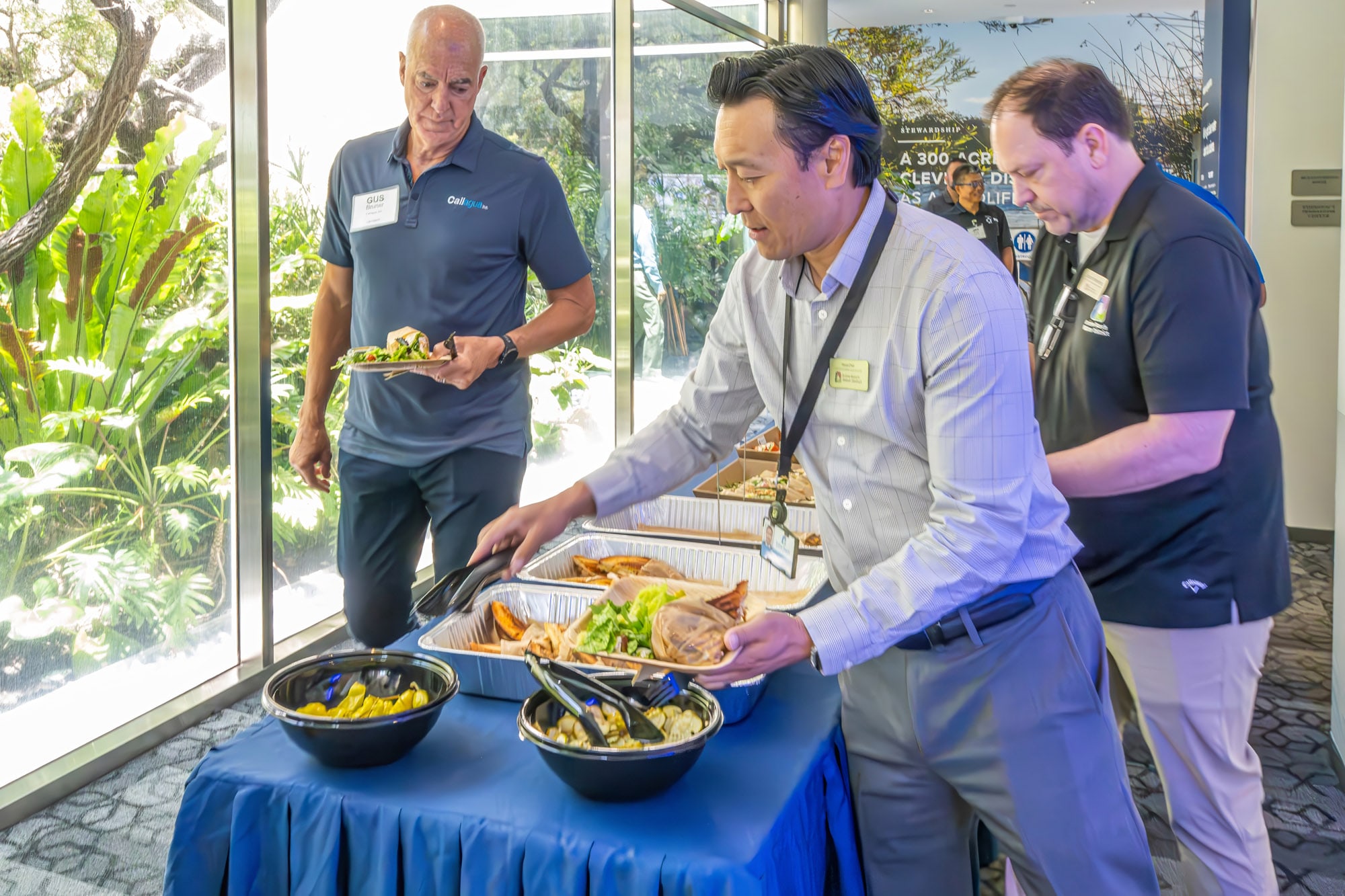 Three men at a buffet table serving themselves from metal trays in a bright indoor event space.