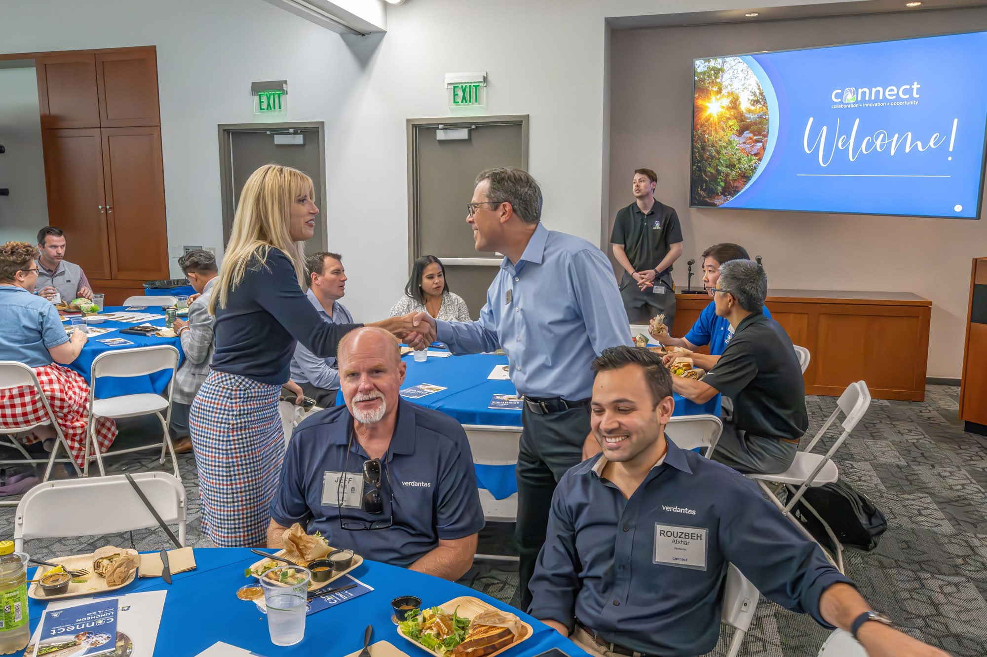 Two professionals shake hands at a networking event in a conference room, with a welcome slide 'Welcome' in the background.