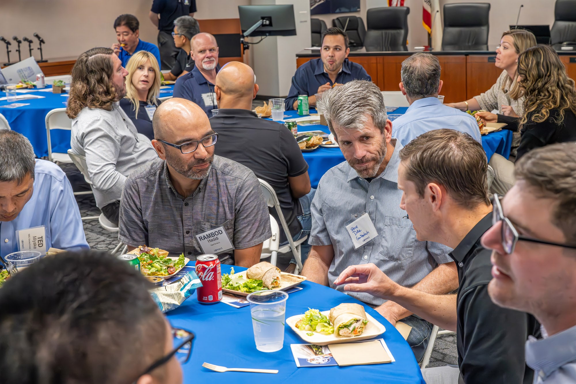 Group of professionals seated around blue-tablecloth tables, sharing lunch at a daytime conference.