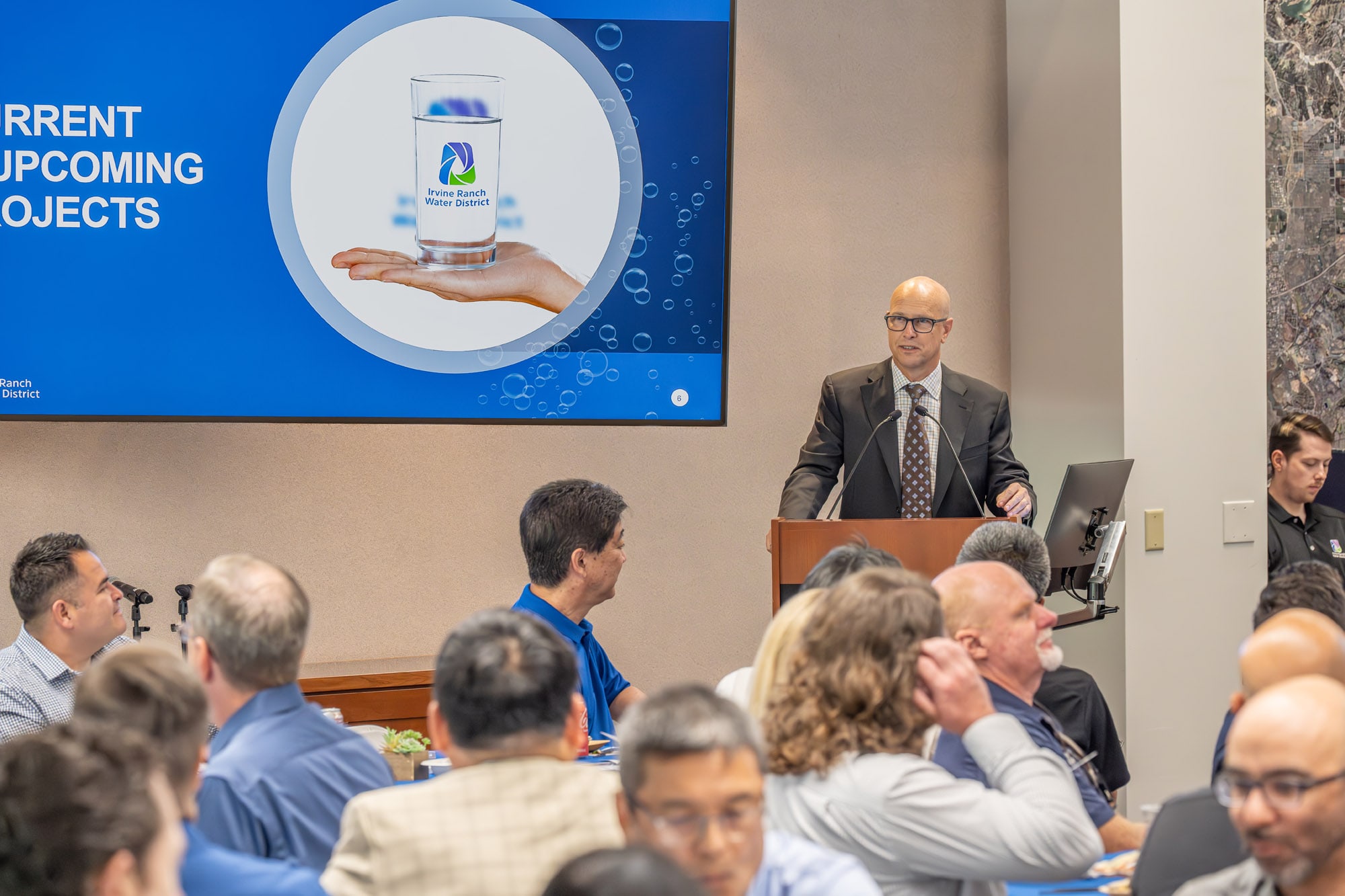 Speaker at podium presenting to audience with a blue slide showing a glass of water and Irvine Ranch Water District logo.