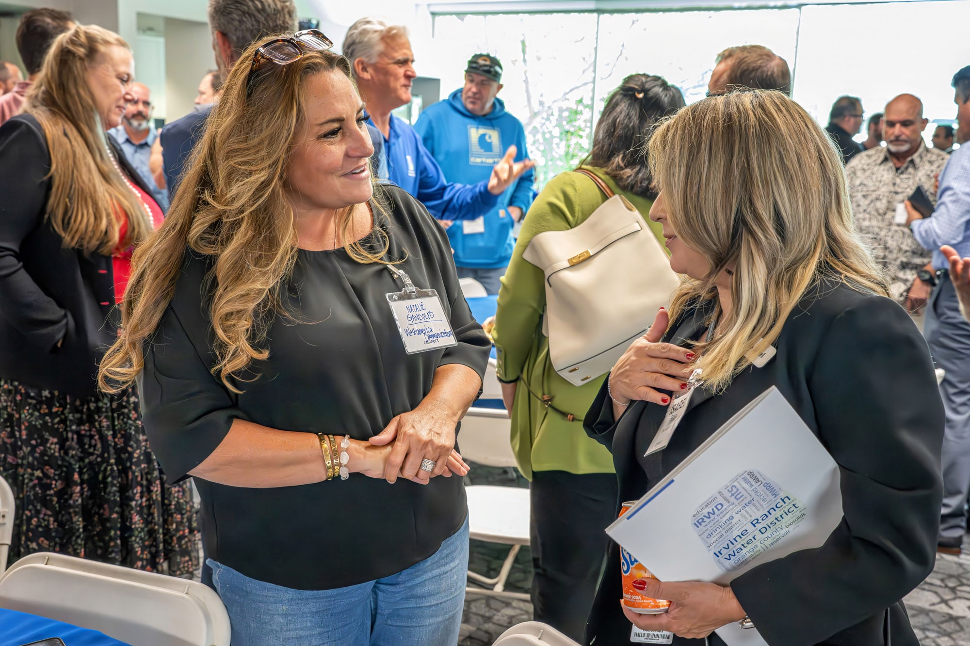 Two women in a crowded conference room chat, with name badges and diverse attendees in the background.