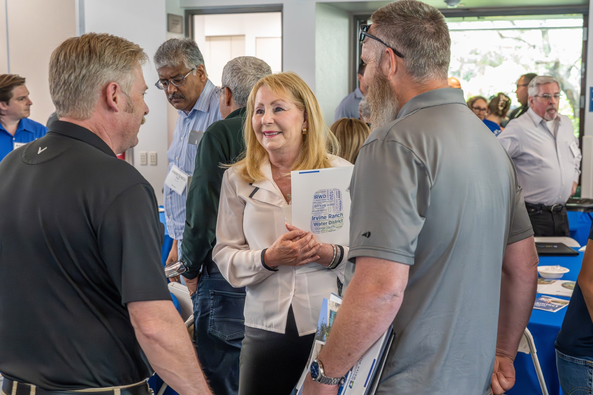 Smiling woman in a light beige blazer engages with attendees at a networking event, holding an Irvine Ranch WD brochure.