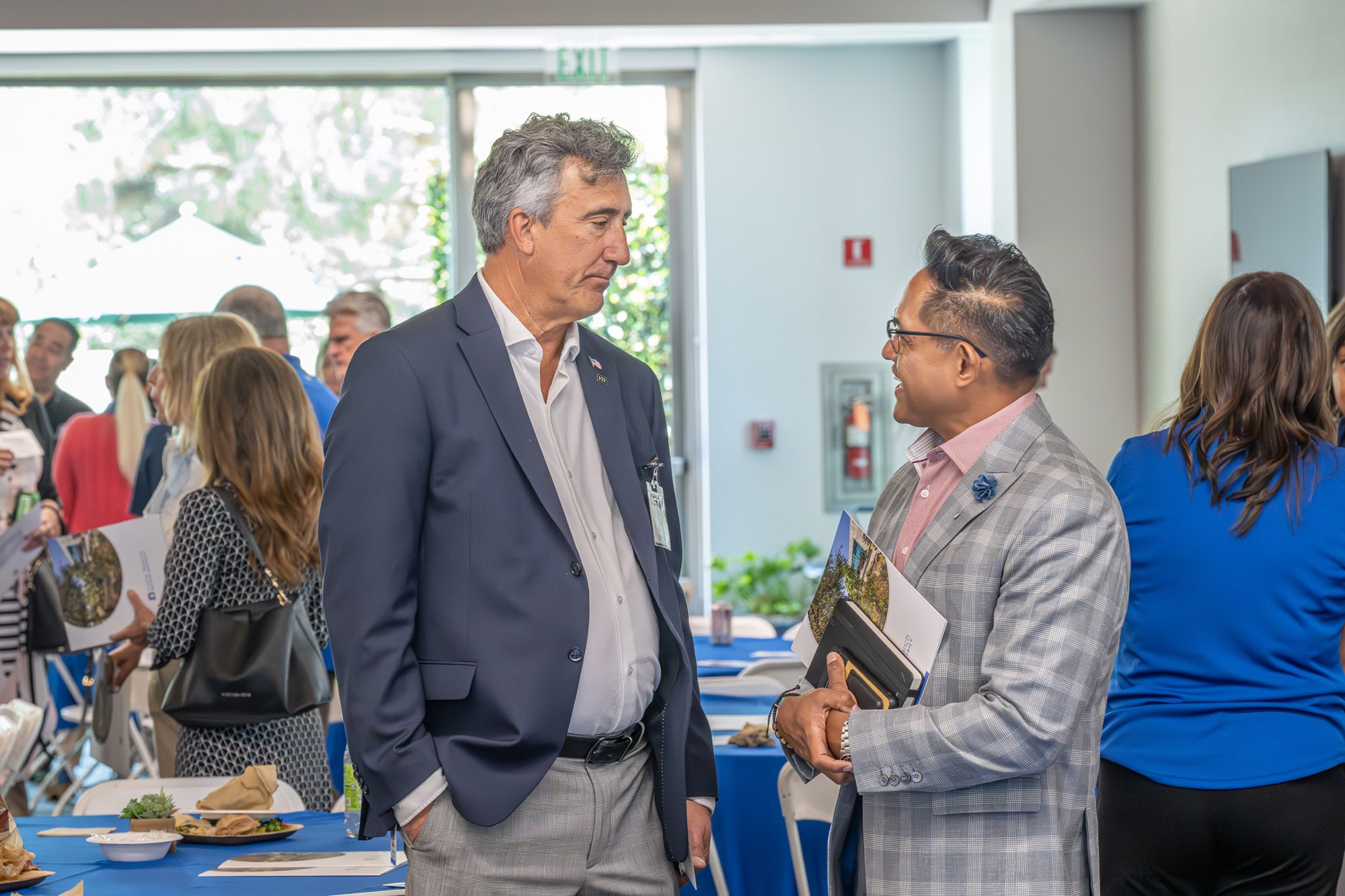 Two professionals in suits converse at a networking event, with blue-table reception setup and attendees in the background.