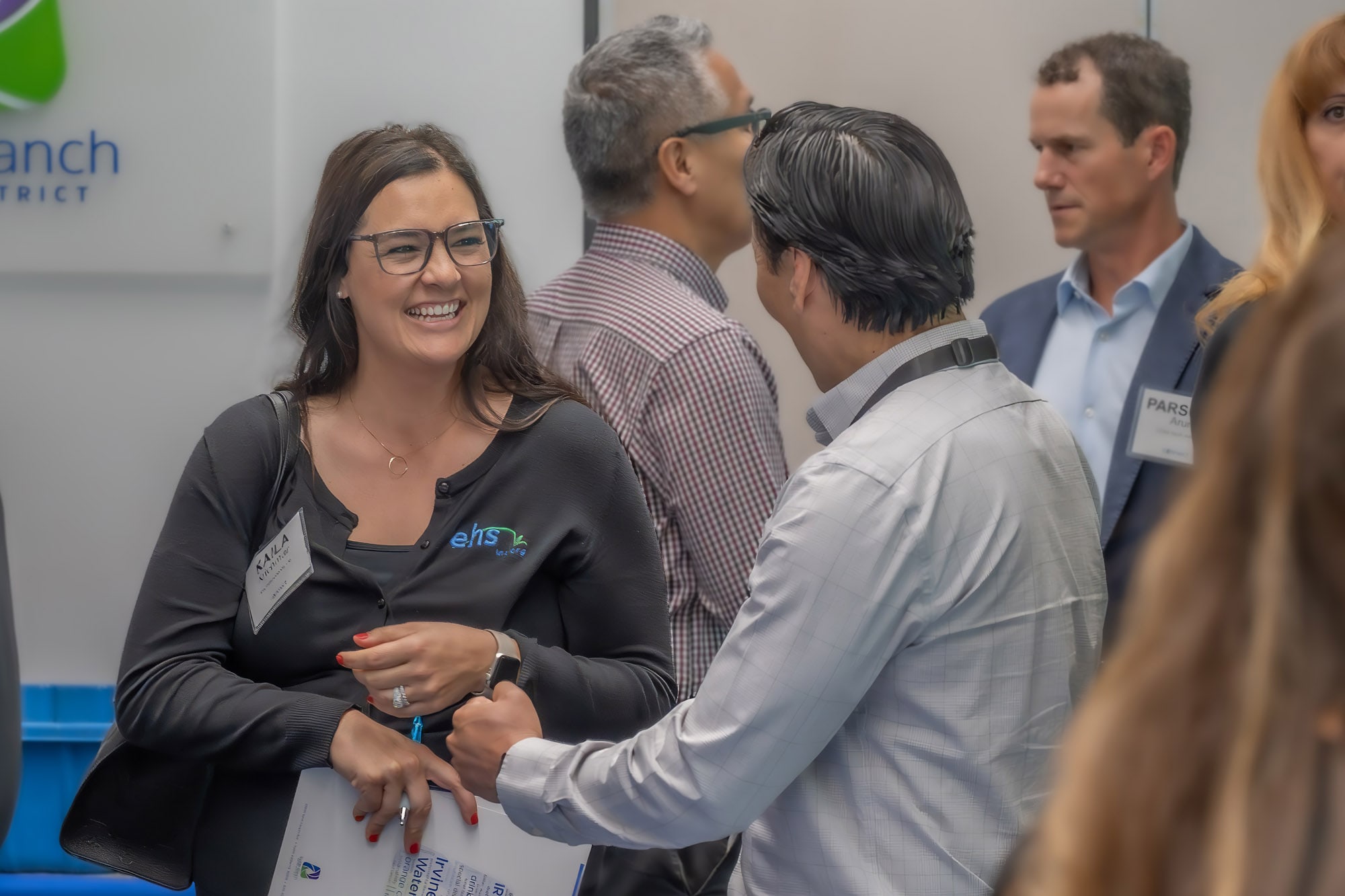 Smiling woman with glasses in a dark cardigan chats with a man in a light shirt at a conference.