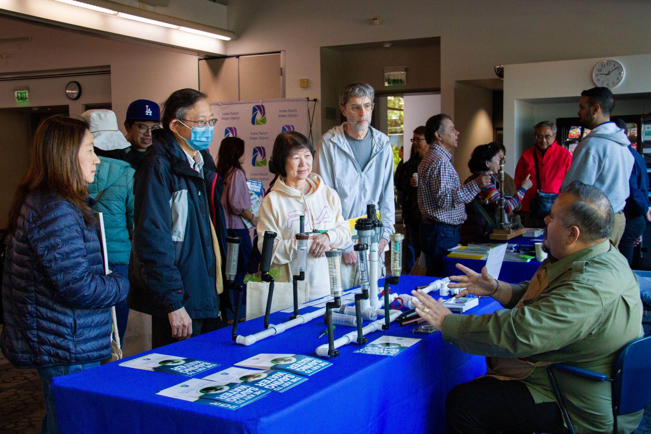 Crowd at an indoor expo watches a plumbing demonstration with PVC pipe rigs on a blue table.