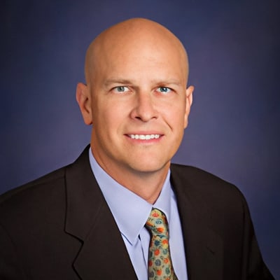 Bald man in a dark suit, light blue shirt, and patterned tie, posing for a professional headshot against a blue backdrop.