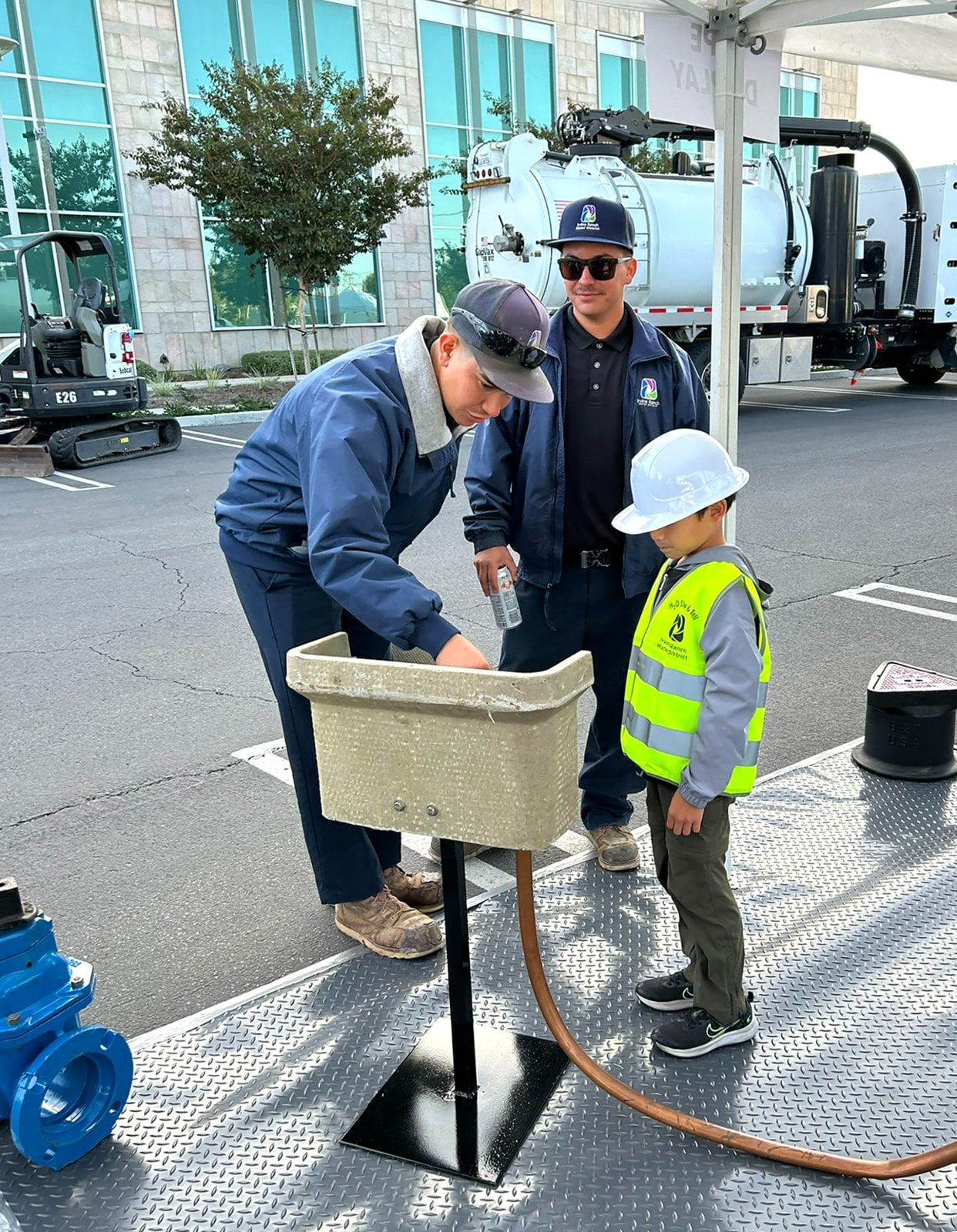Workers perform a sample collection demo with a beige trough and hose while a child in a hi-vis vest watches.
