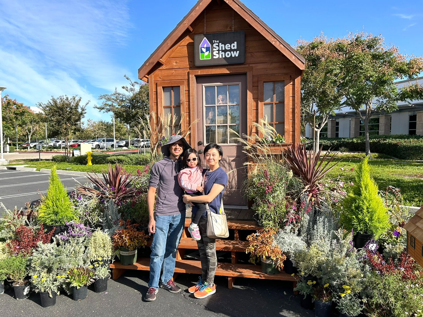 Family posing in front of a wooden shed display labeled The Shed Show, surrounded by colorful potted plants.