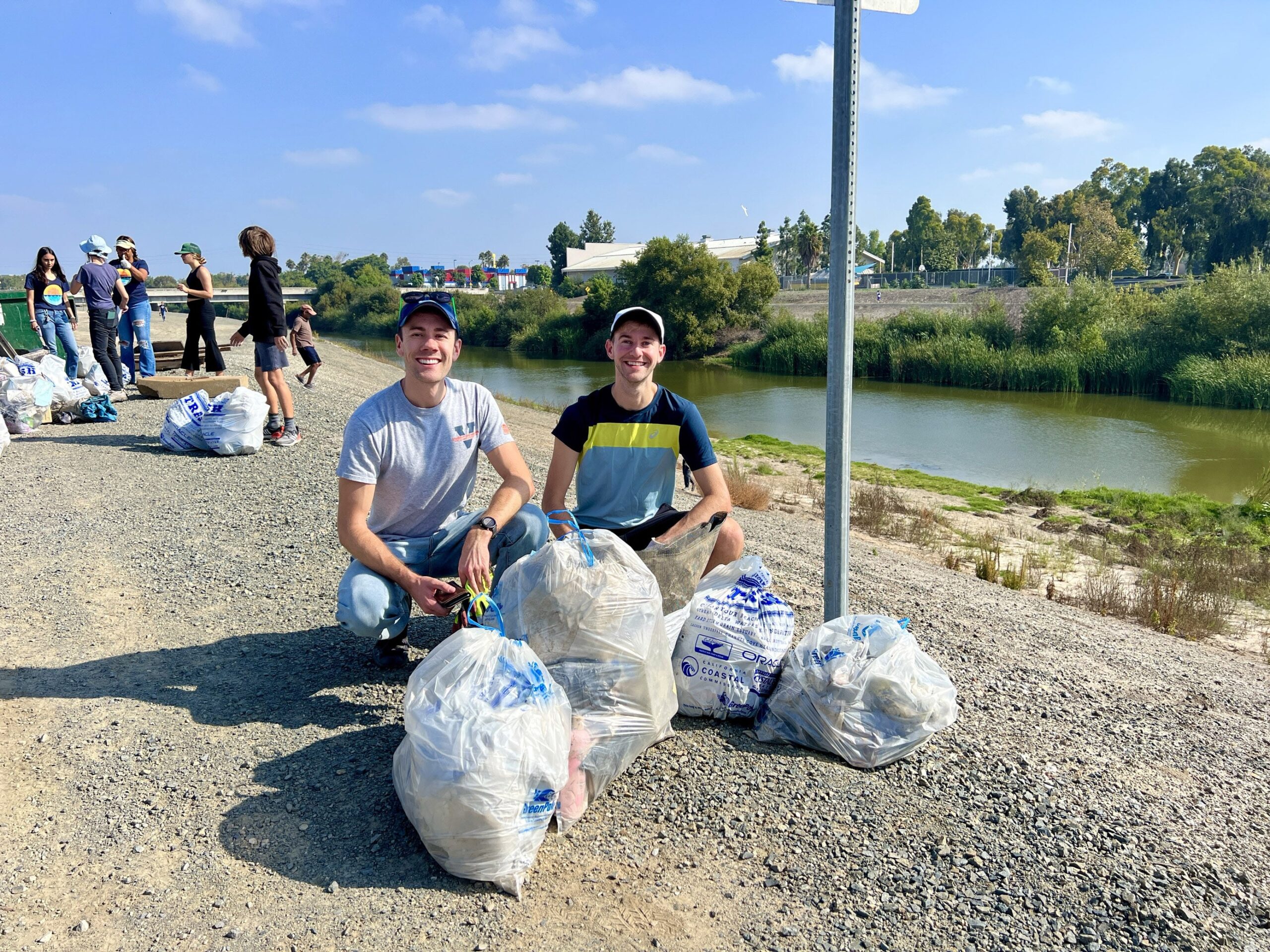 Two men kneel beside filled trash bags on a gravel riverbank while volunteers clean up nearby.