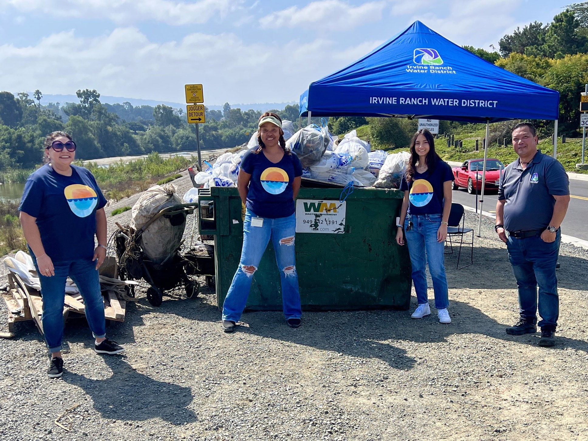 Four volunteers in navy shirts with a sunset logo pose beside a full dumpster under a blue Irvine Ranch Water District canopy.