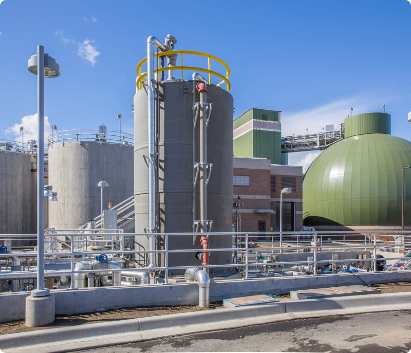 Industrial plant with large cylindrical tanks, pipes, and a green dome building under a clear blue sky.