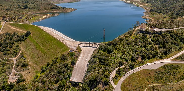Aerial view of a dam forming a reservoir, surrounded by green hills and winding roads.