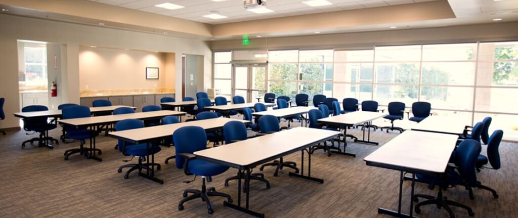 Bright classroom with blue chairs and white tables arranged in rows beside large windows.