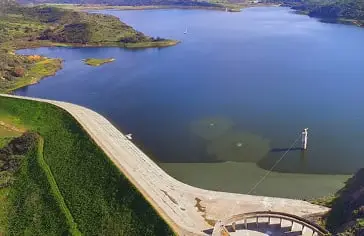 Curved spillway along a dam beside a large blue reservoir with green hills.