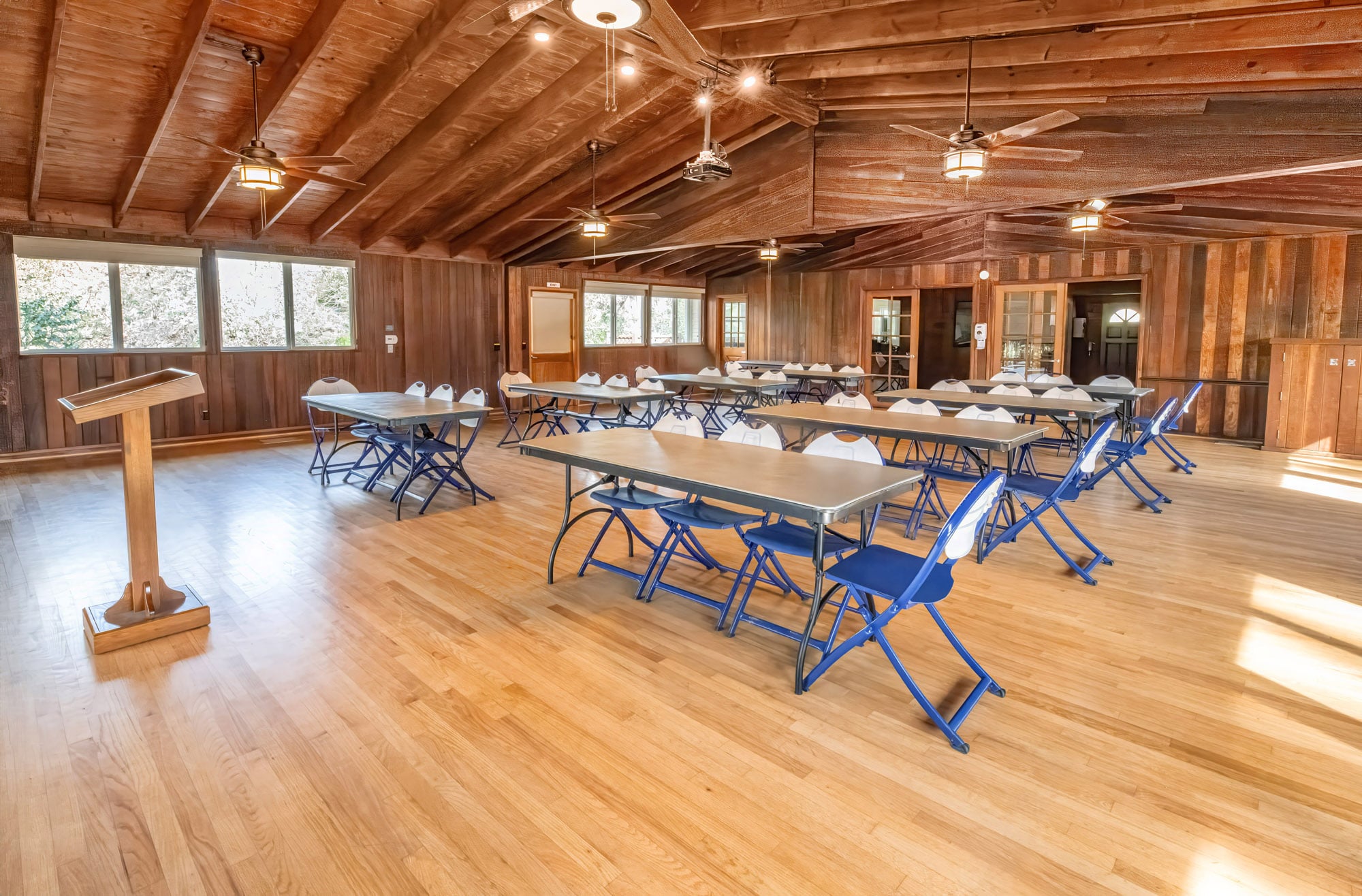 Wood-paneled event hall with long tables and blue folding chairs, a podium, and ceiling fans.