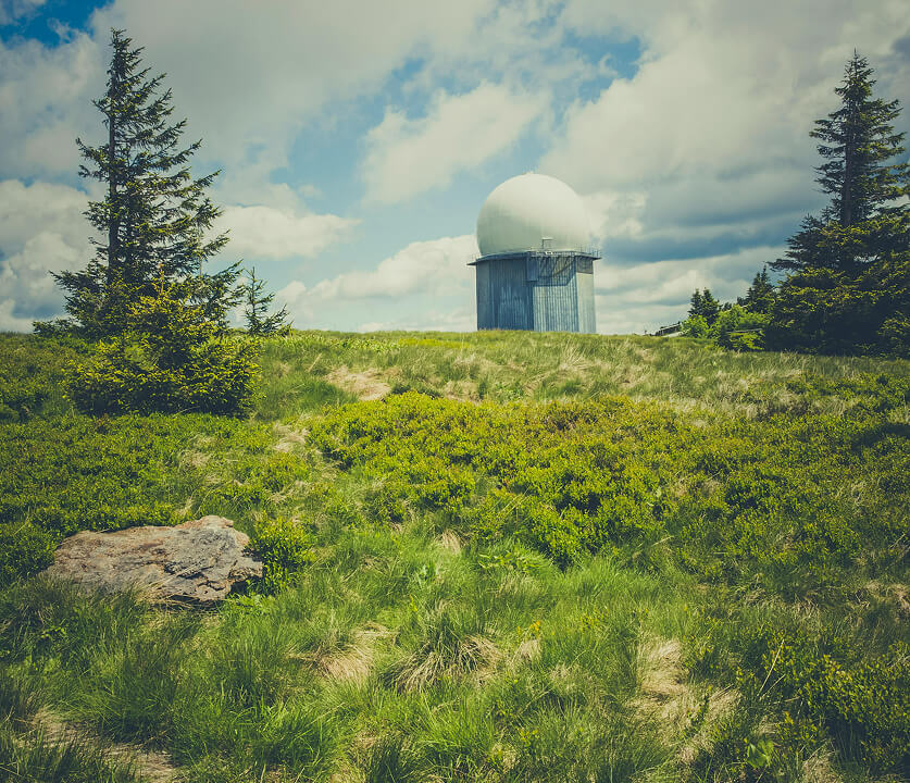 Radar dome atop a small metal building on a grassy hill, framed by evergreen trees under a partly cloudy blue sky