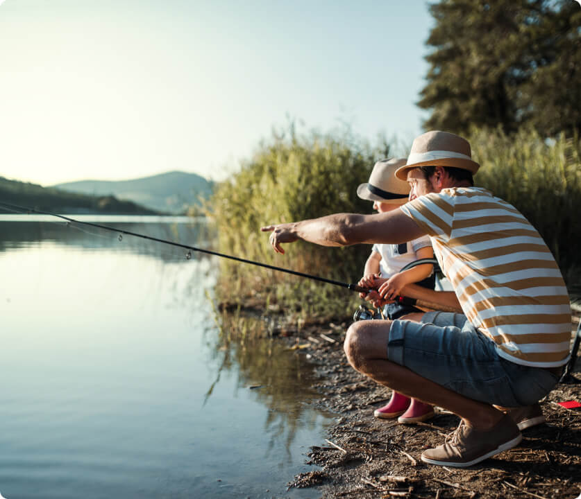 Two people fishing by a calm lake; a man in a striped shirt crouches and points as a child watches.