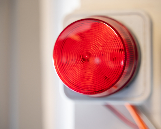 Red circular emergency alarm light mounted on a white wall panel with visible wiring.
