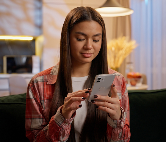 Woman with long dark hair wearing a red plaid shirt sits on a sofa and looks at her smartphone