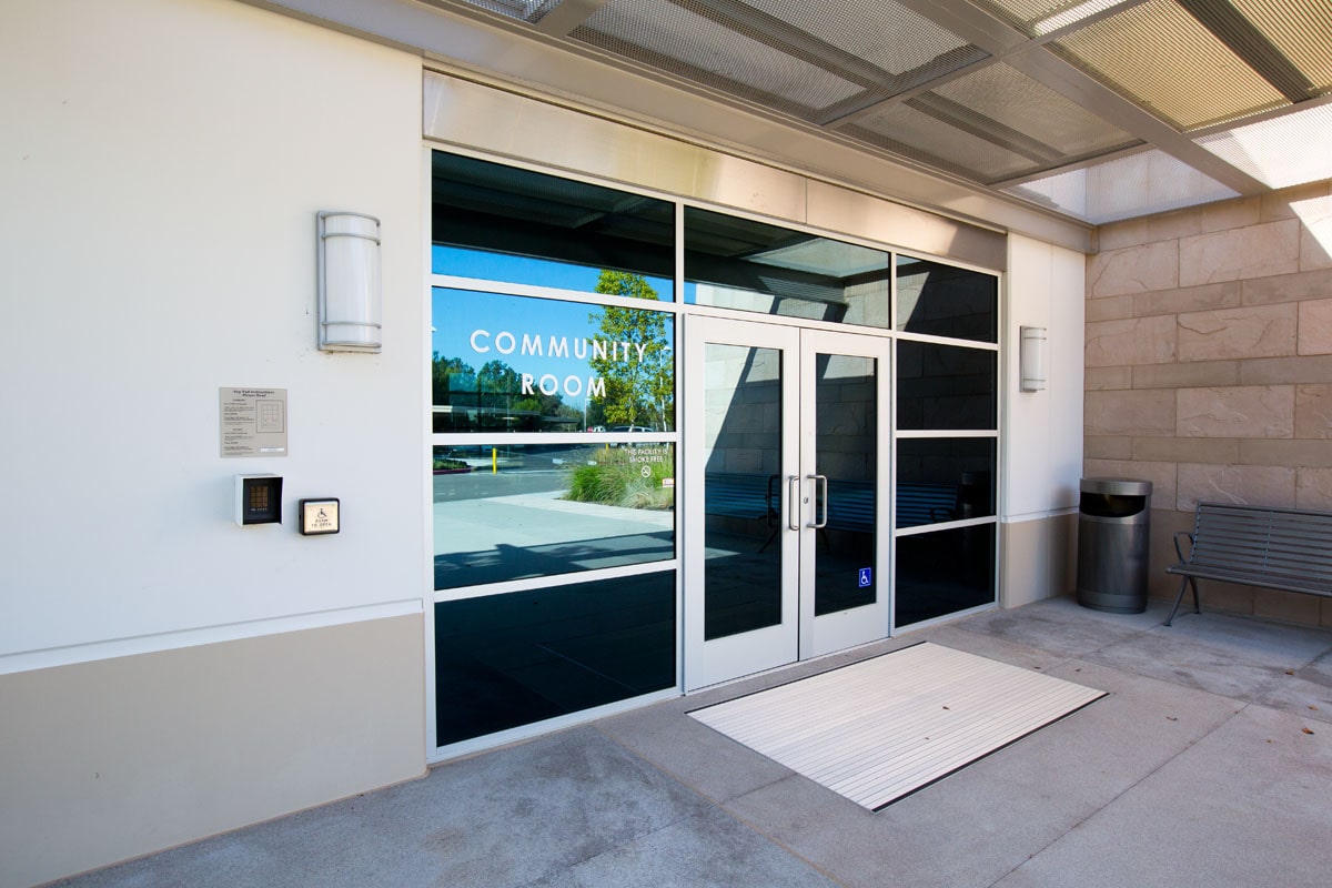 Glass double doors labeled Community Room with wheelchair accessibility sign, bench to the right, and a ramp mat outside.