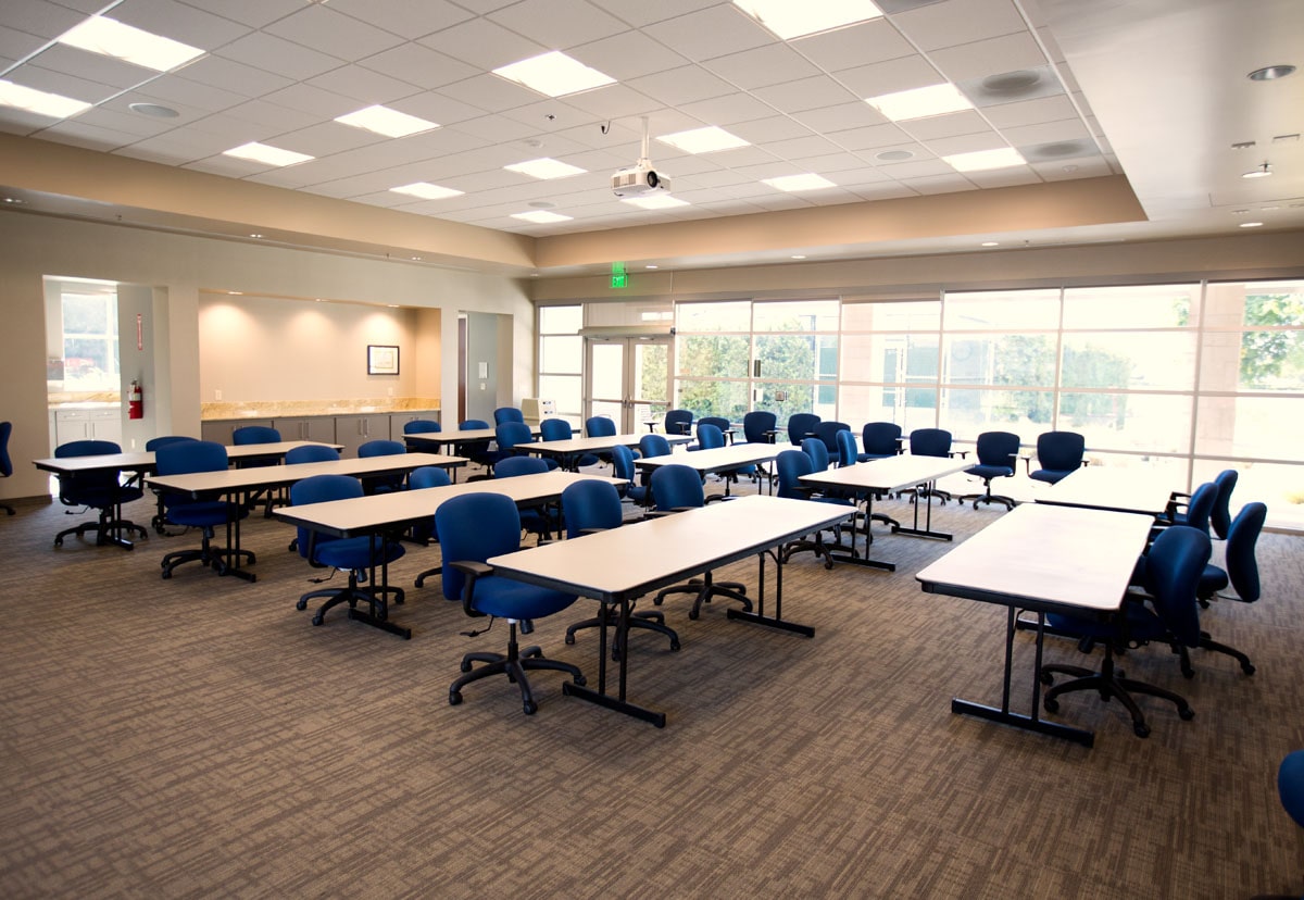 Conference room with long white tables and blue rolling chairs arranged in rows, large windows at the back.