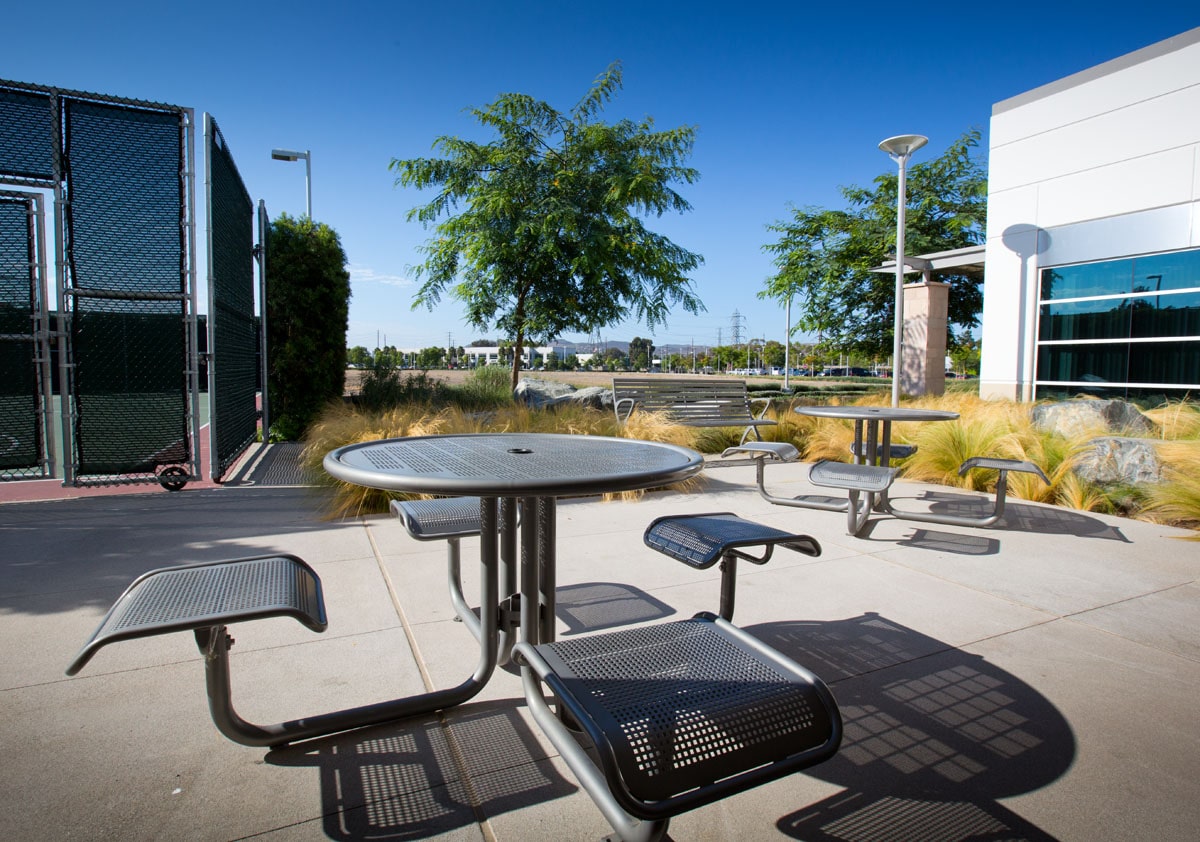 Outdoor metal picnic tables and benches on a paved plaza with trees, grasses, and a modern building nearby.