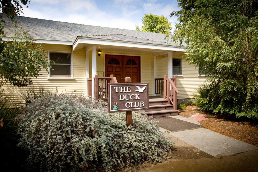 Front view of a pale-yellow cottage with a wooden porch and a sign that reads The Duck Club in front of evergreen shrubs.