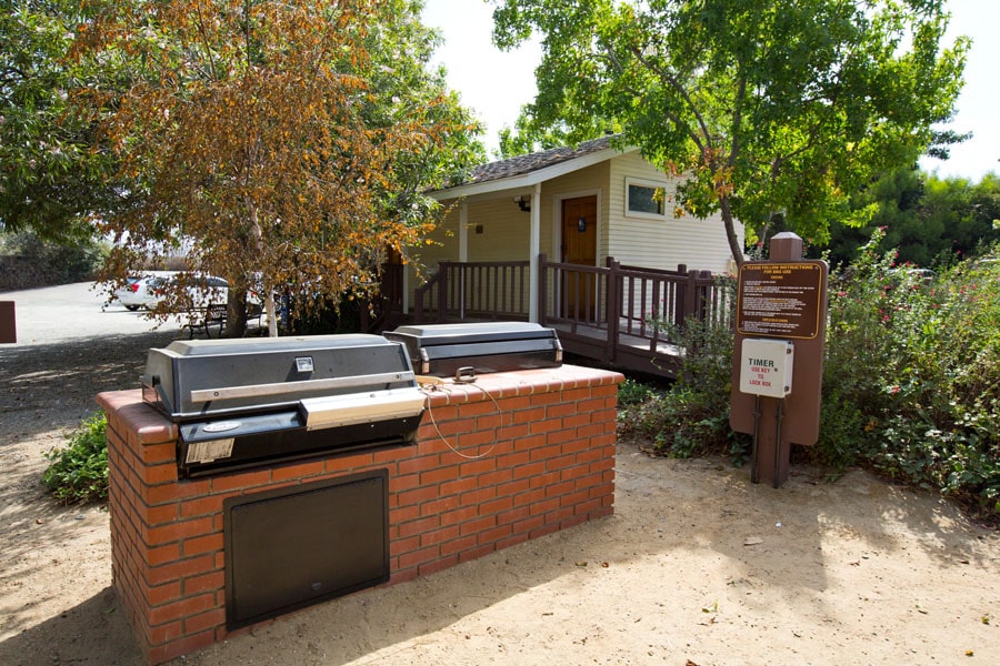 Two charcoal grills built into a brick counter in a park picnic area, with a small beige building and trees in the background.