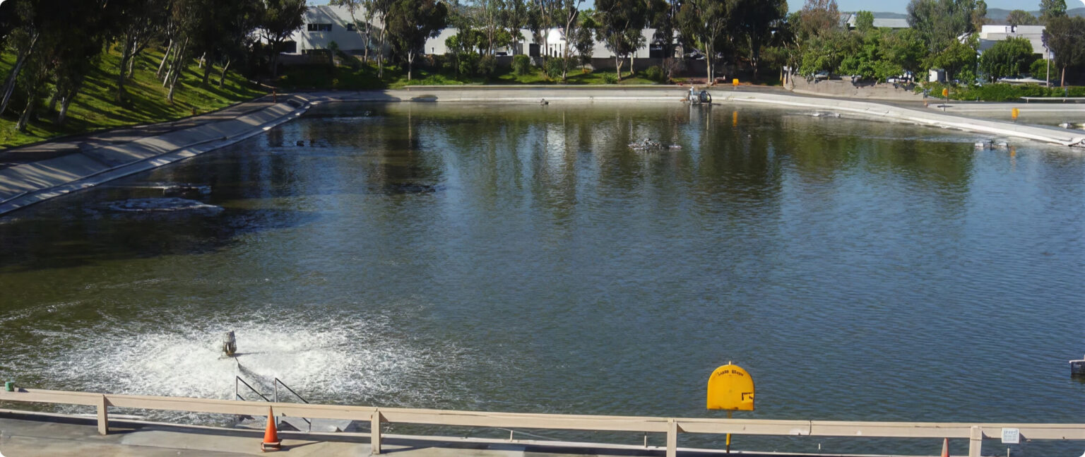 Rectangular urban water basin with concrete banks, grassy slopes, and trees; a yellow buoy sits in the foreground.