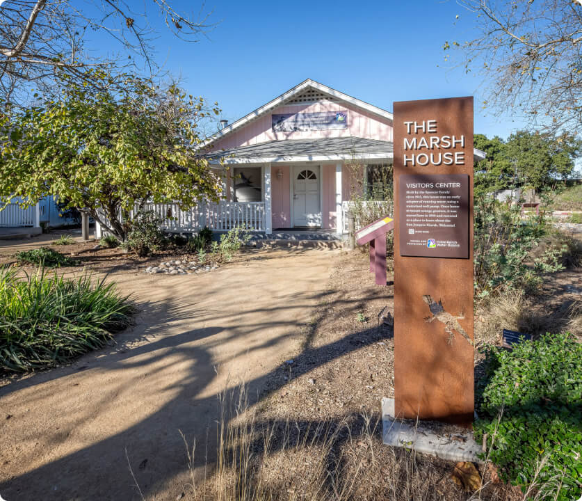 Pink cottage-style building with white trim and a front porch, and a tall rust-colored sign reading The Marsh House Visitors Center.