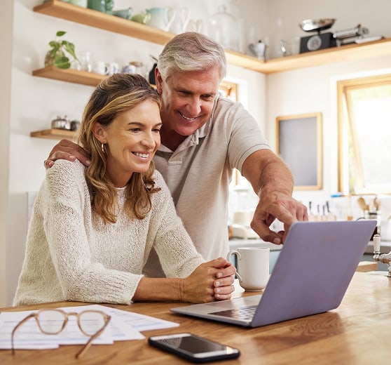 Older couple using a laptop at a kitchen island, smiling and pointing at the screen.