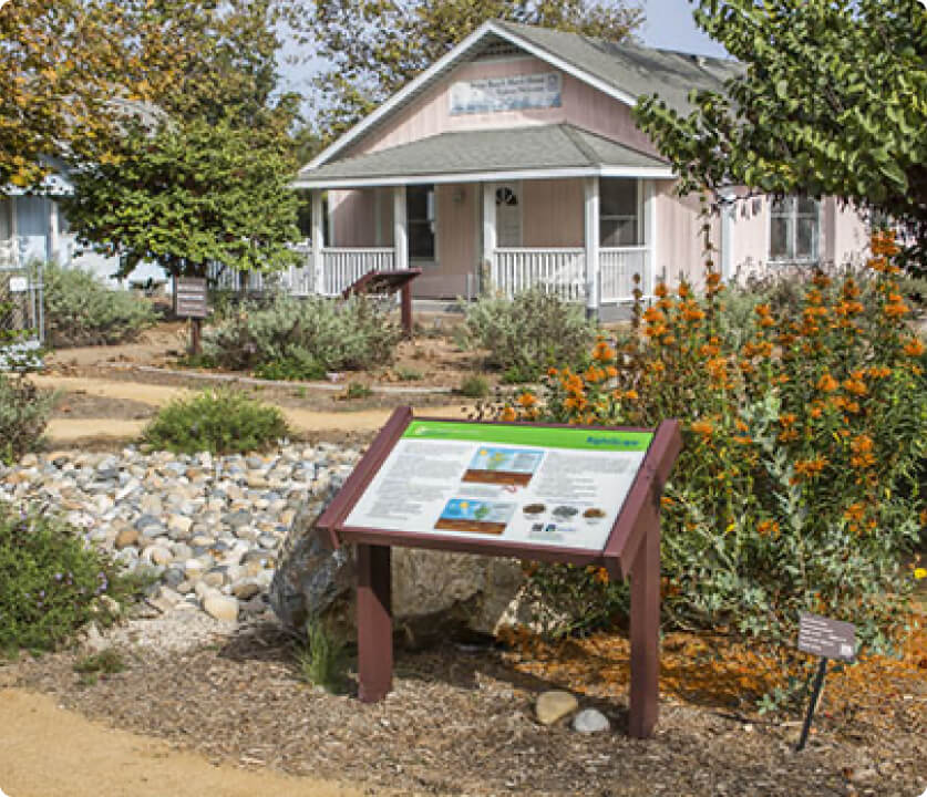 Pink single-story cottage with a front porch, amid a drought-tolerant garden and an information display.