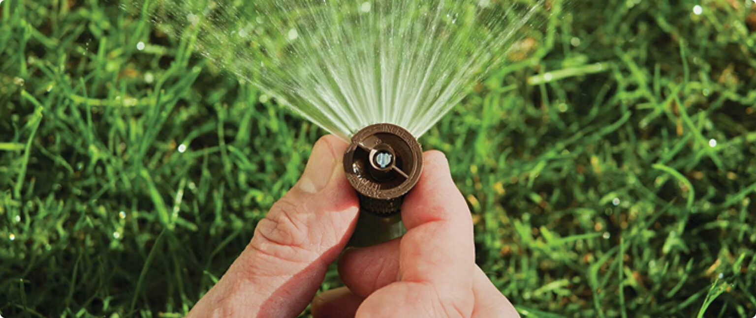 Hands holding a small irrigation sprinkler spraying water across a green lawn.