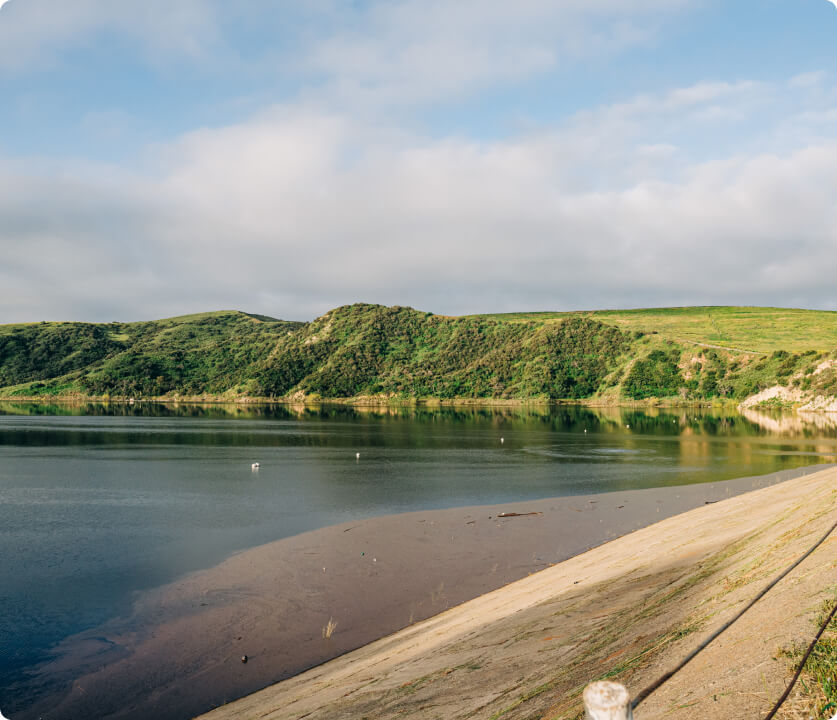 Calm lake lined with green hills, a sandy shoreline, and a concrete promenade along the water.