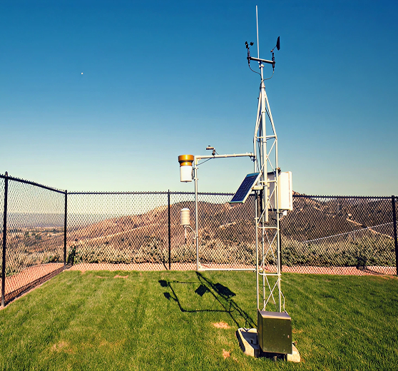 Weather station with solar panel, wind vane, and sensors on a grassy fenced area overlooking hills.