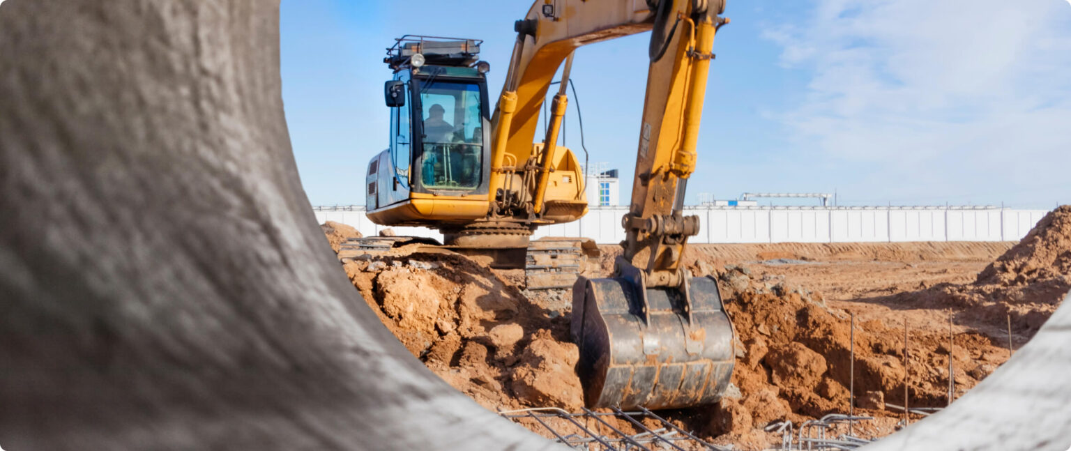 Yellow excavator at a dirt construction site, framed by large concrete pipes in the foreground.