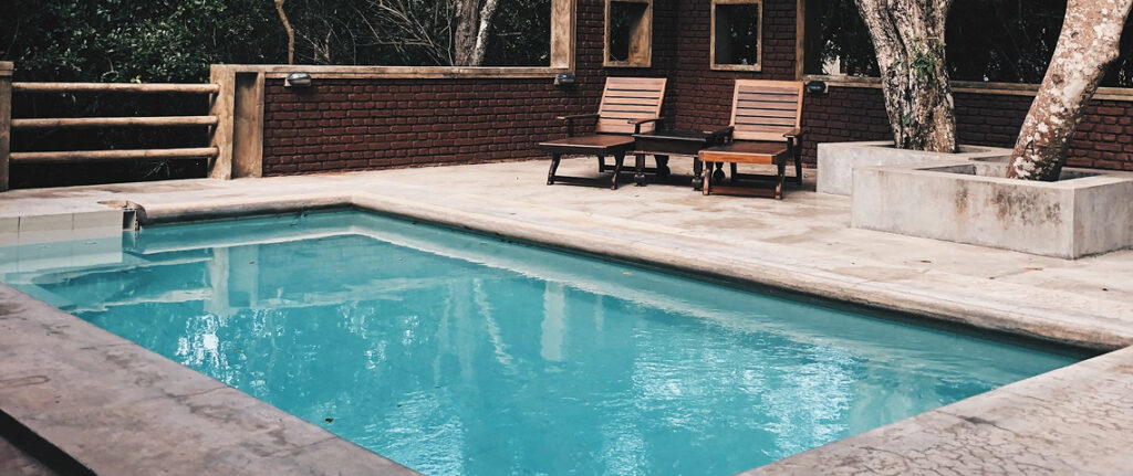 Outdoor rectangular pool with turquoise water, concrete deck, and wooden lounge chairs beside a brick wall and trees.