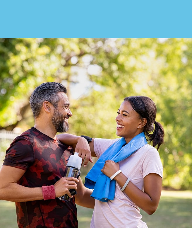 Two fit adults chat and smile after a workout in a sunny park; the man holds a water bottle and the woman wears a blue towel around her neck.